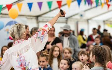 Frau von hinten mit bunter Bluse reckt Arm mit Mikro in die Höhe und singt mit Kindern vor ihr