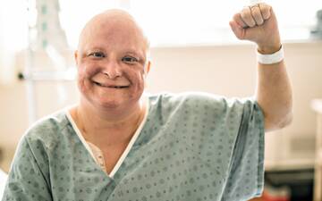 Patient ohne Haare im OP-Kittel reckt die Hand zur Faust und lacht