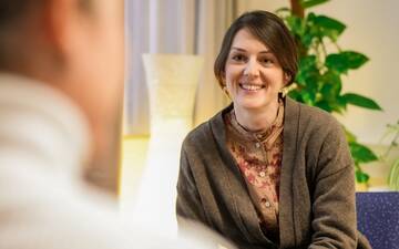 Freundliche, junge Frau mit brauner Weste berät eine Person, im Hintergrund eine Leuchte und eine Zimmerpflanze