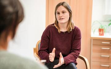 Junge Frau mit dunkelrotem Pullover erklärt einer anderen Person etwas