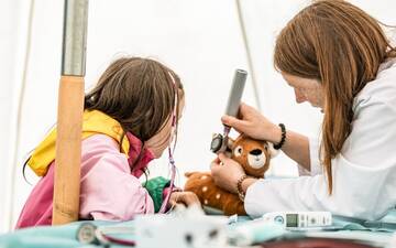 Frau simuliert Ohruntersuchung an einem Stofftier, Kind schaut interessiert zu