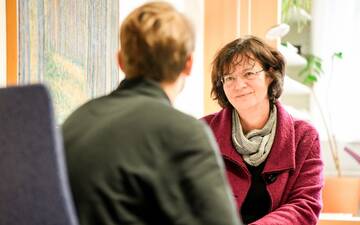 Freundliche Frau mit roter Weste berät eine Person, im Hintergrund eine Zimmerpflanze