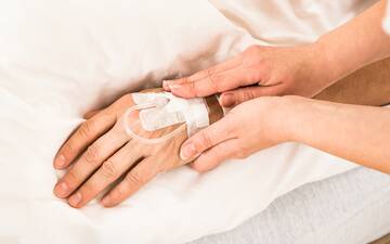 Hand mit Kanüle liegt auf einer Bettdecke, zwei andere Hände halten die Hand