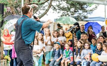 Mann mit Latzhose von hinten auf einer Bühne führt Kindern etwas vor