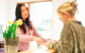 Rezeption einer Praxis, im Vordergrund links scharf abgebildet ein Topf mit Osterglocken, weiter hinten unscharf zwei Frauen