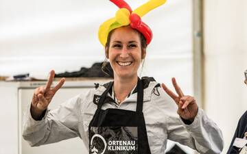 Junge, lachende Frau mit Schürze mit dem Ortenau Klinikum Logo und einem Hut aus Luftballonschlangen in rot und gelb